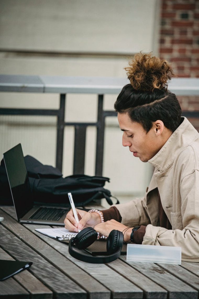 young serious ethnic man with trendy hairstyle writing at table